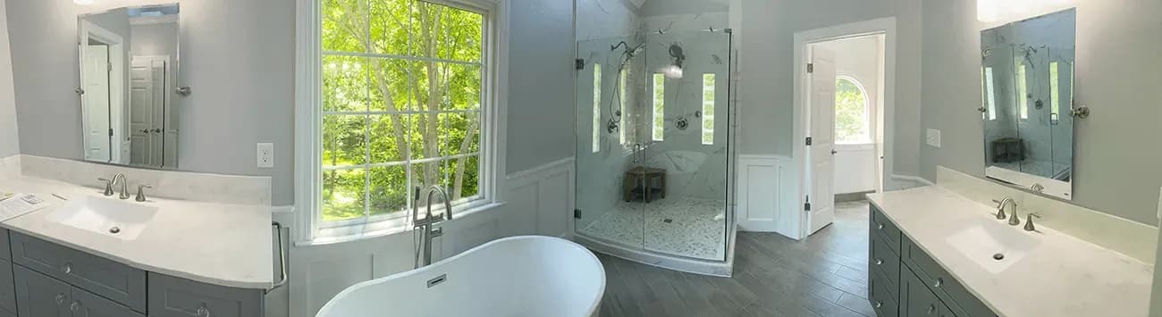 A panoramic view of a newly-remodeled bathroom with two vanities, white countertops, gray cabinets, a walk-in shower, and a large free-standing soaking bathtub.
